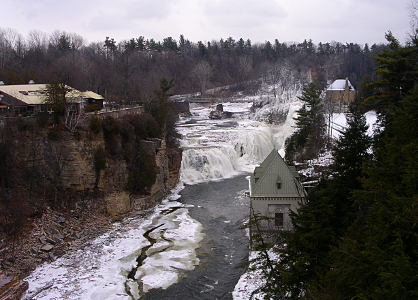 [The left side of the image is a rocky cliff leading to the snow-covered water. The waterfalls are at the back of the image with rapids just before the falls. On teh right side is a two story white building with the second story being under a slanted roof.]